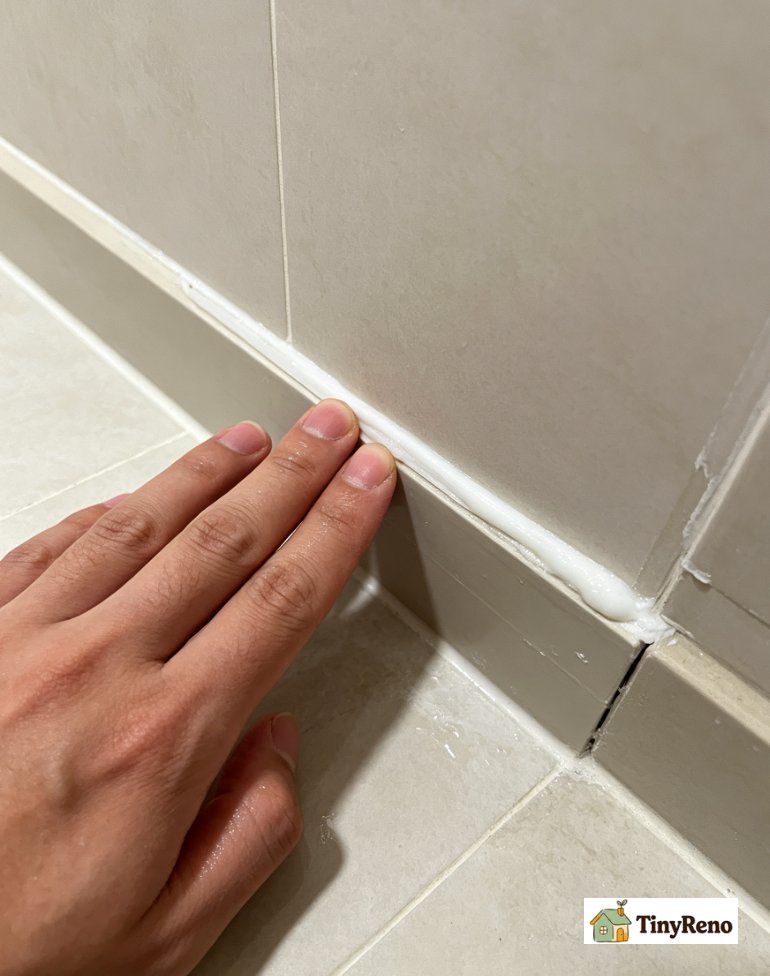 Renter applying caulk to baseboard-floor gap, bathtub tile seam, trim corner joint, window frame gap, countertop backsplash seam, smoothing bead with wet finger.